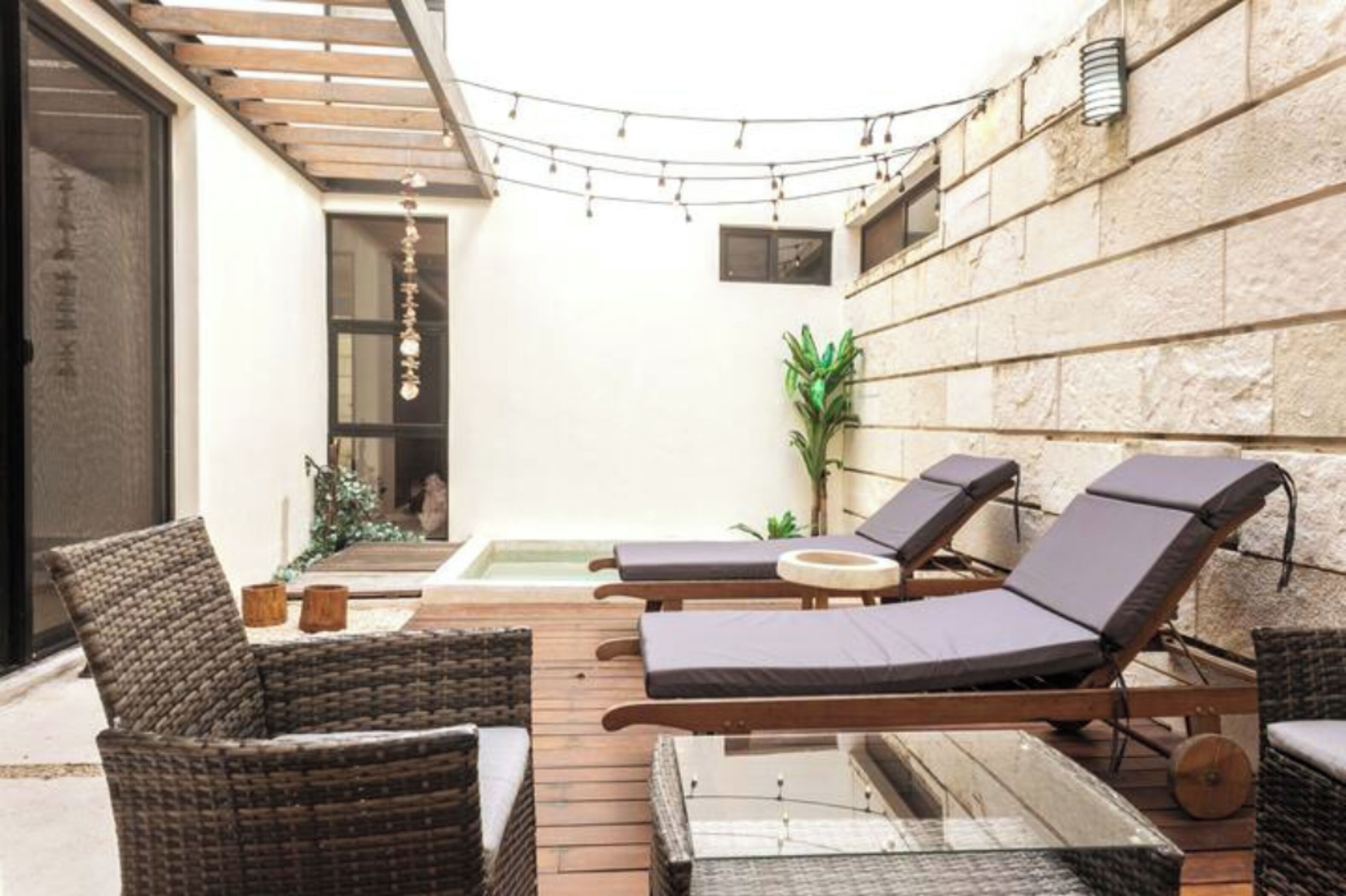 A sunlit outdoor seating area with wicker furniture and a small table, a stone wall, and a string of lights.