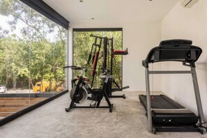 A gym with a treadmill, weight machine, and a large window offering a view of trees and a yellow vehicle outside. - Magnificent Apartment Pool Gym (Tulum)