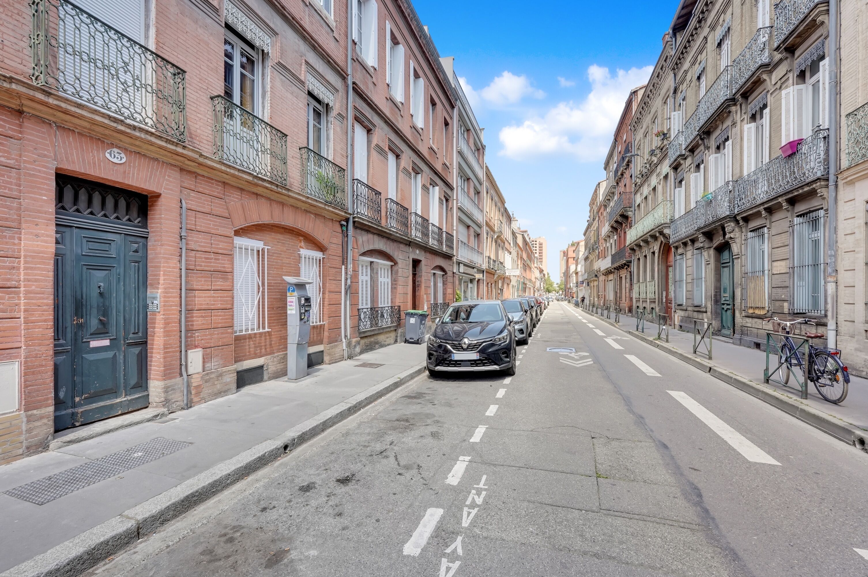 Una calle estrecha bordeada por edificios de varios pisos, con balcones de barandilla metálica, un automóvil estacionado y una bicicleta.
