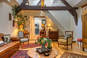 A cozy living room with a high ceiling, wooden beams, a sofa, chairs, a coffee table, and a staircase leading to another room.