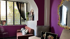 Ein Zimmer mit lila Wand, einer Couch, einem Bett, einem Spiegel und einem Fenster mit Vorhängen.