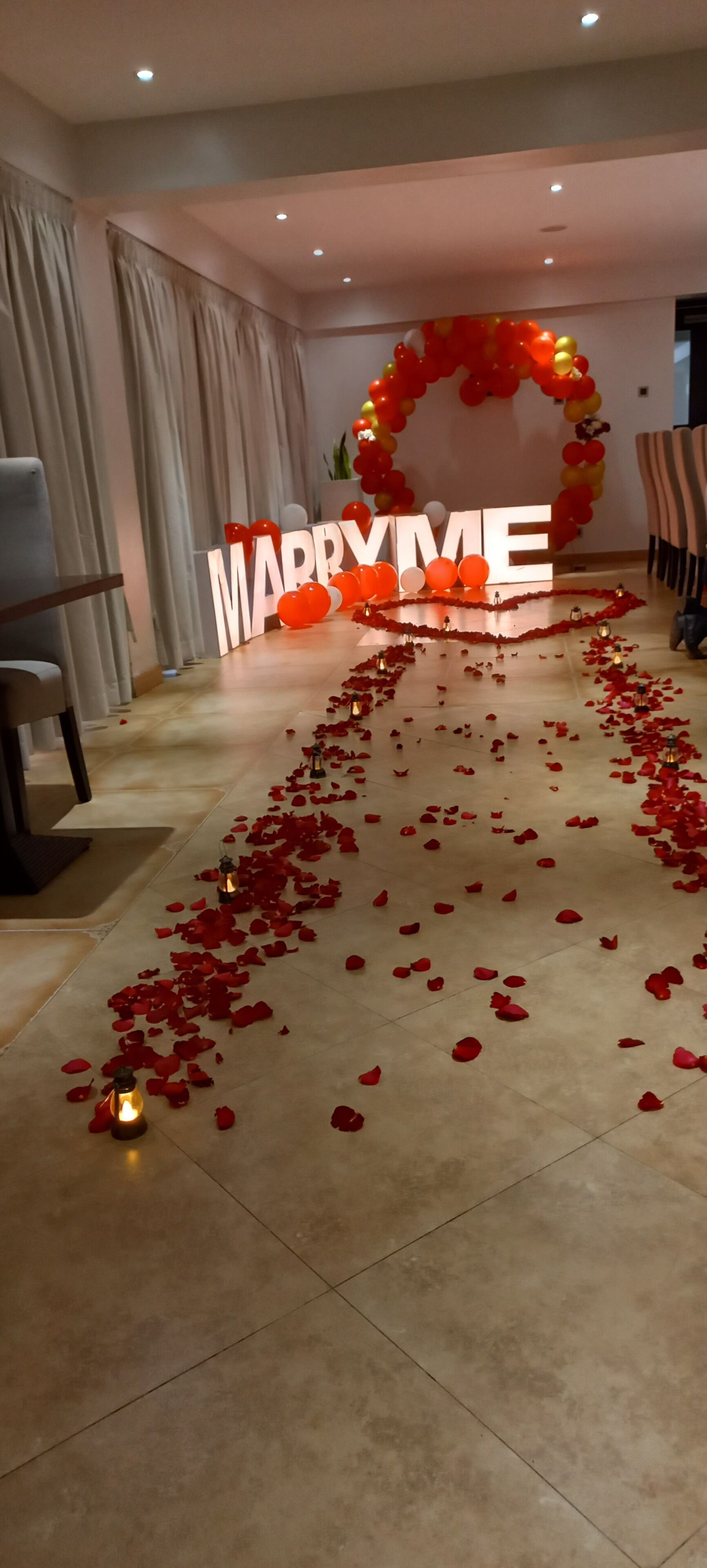 Decoración para una boda con un cartel que dice 'MARRY ME', globos rojos y pétalos de rosa en el suelo.