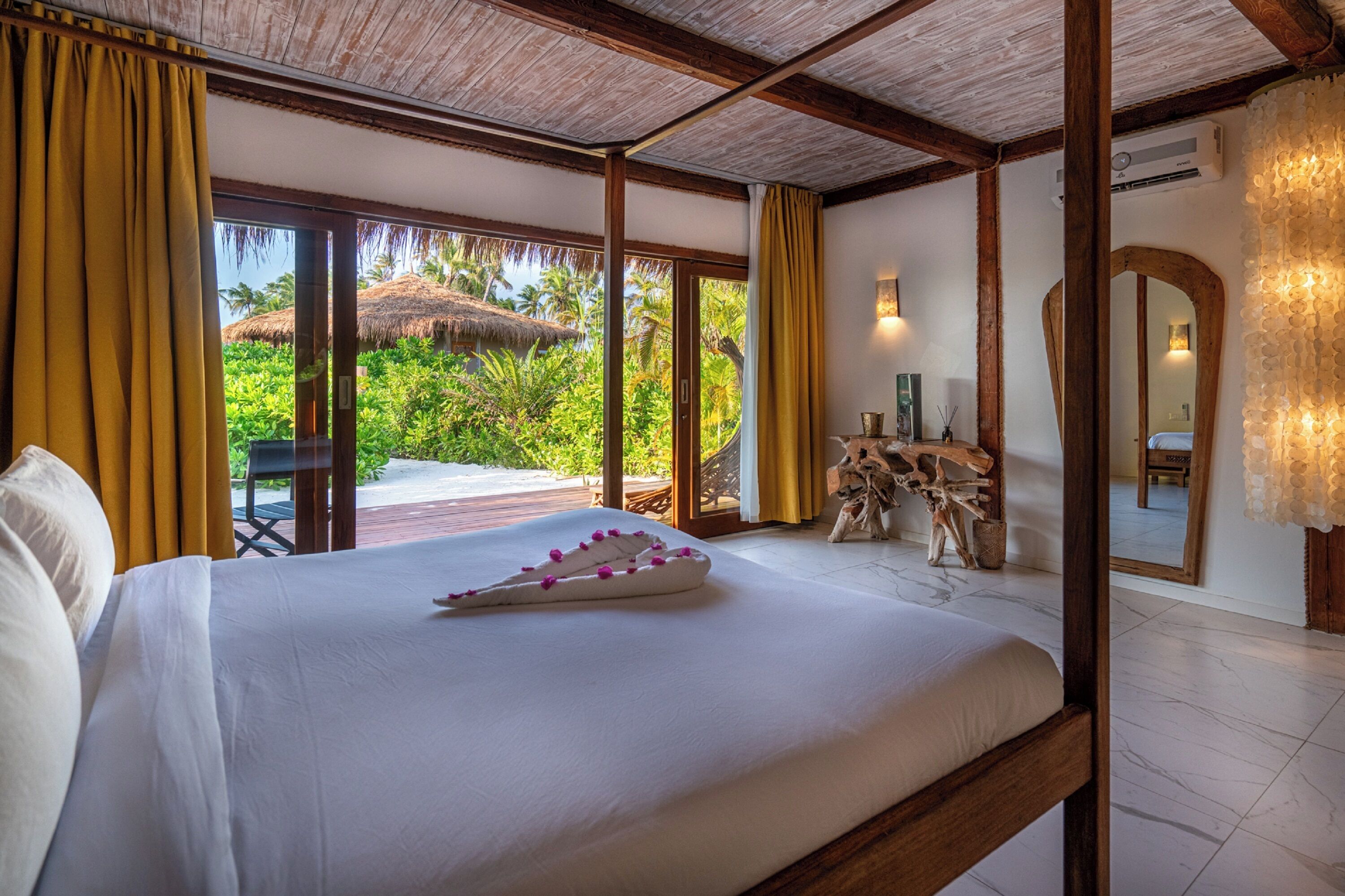 A bedroom with a large bed, a view of tropical vegetation, and a wooden ceiling.