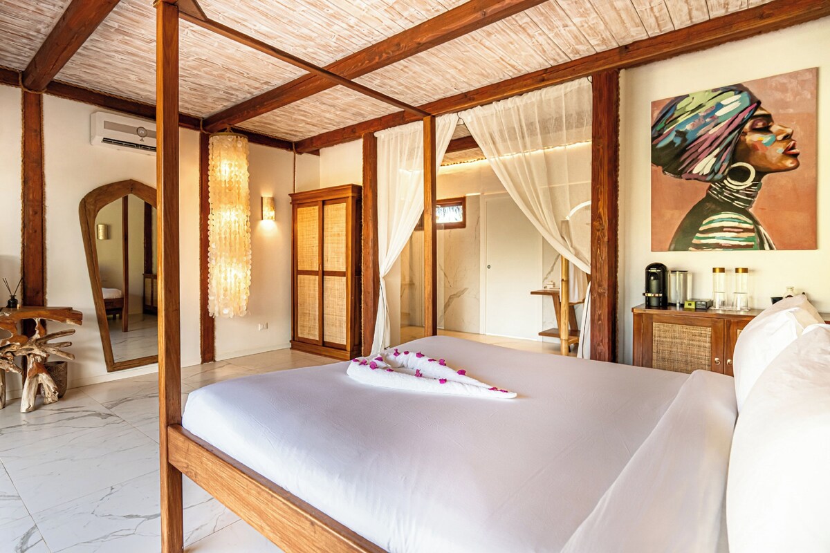 A bedroom with a large bed, wooden beams, a mirror, a painting, and a bedside table with a lamp.