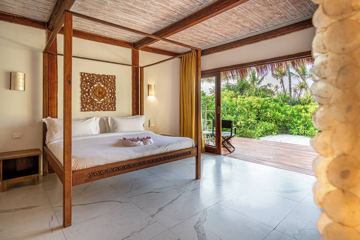 Una cama con dosel de madera y ropa blanca, un adorno en la pared encima y vistas de vegetación exuberante a través de un ventanal.