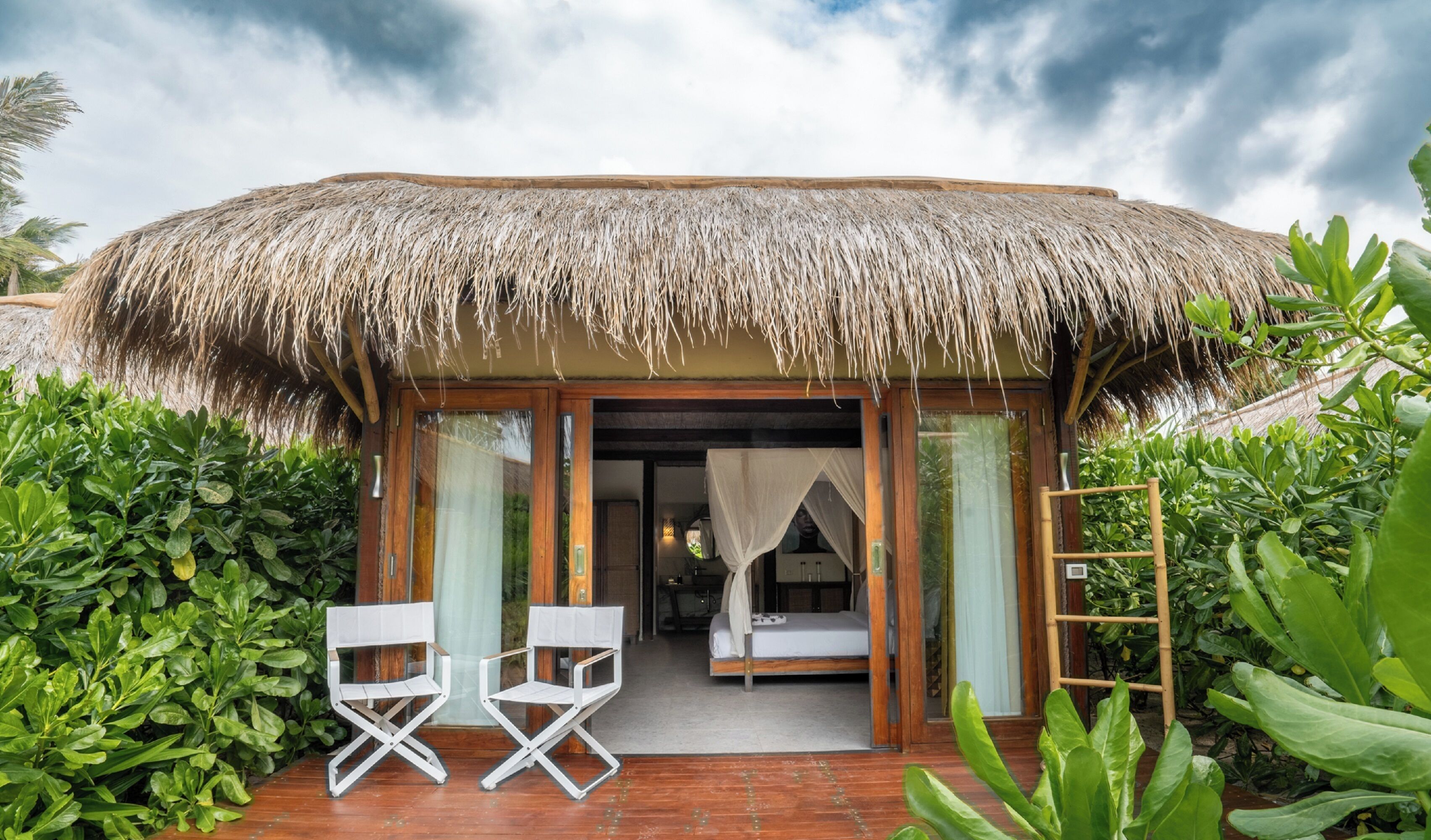 Una cabaña con techo de paja y terraza de madera, con dos sillas blancas y una cama con dosel.