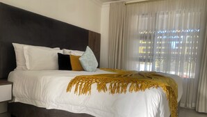 Una cama con colcha blanca y dorada, cabecera oscura y ventana con cortinas traslúcidas.