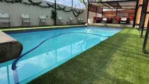 Una piscina con césped artificial, sillas blancas y una zona de estar cubierta con mesas.