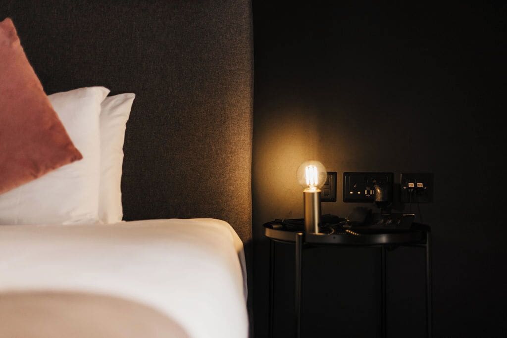 A hotel room with a bed, a nightstand with a lamp, and a control panel.