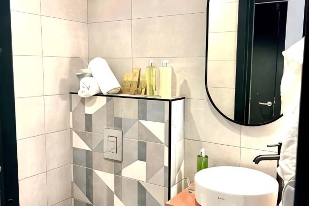 A bathroom with a modern design, featuring a geometric tile pattern on the walls, a glass shelf with toiletries, a white sink, and a round mirror.