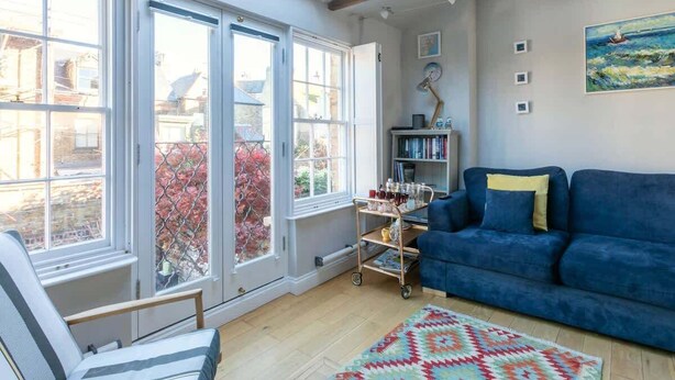 Una habitación con un sofá azul, una silla, una estantería y una ventana con vistas a edificios y árboles.