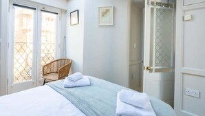 Un dormitorio con una cama, una silla de mimbre, un baño con un espejo y una puerta con un panel de vidrio esmerilado.