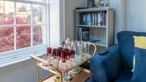 Una habitación con ventana, una estantería, un sofá azul y una bandeja con vasos.