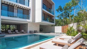A modern two-story building with a swimming pool, lounge chairs, and a view of the surrounding greenery. - Increíble Departamento con Piscina y Gym (Tulum)