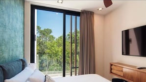 A modern hotel room with a large bed, a flat-screen TV, a wooden nightstand, and a view of trees through a sliding glass door. - Increíble Departamento con Piscina y Gym (Tulum)