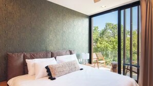 A modern hotel room with a large bed, a view of greenery through floor-to-ceiling windows, and a balcony with a chair and table. - Increíble Departamento con Piscina y Gym (Tulum)