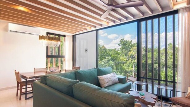 A modern living room with a green sofa, a dining table, and a large window offering a view of trees. - Increíble Departamento con Piscina y Gym (Tulum)