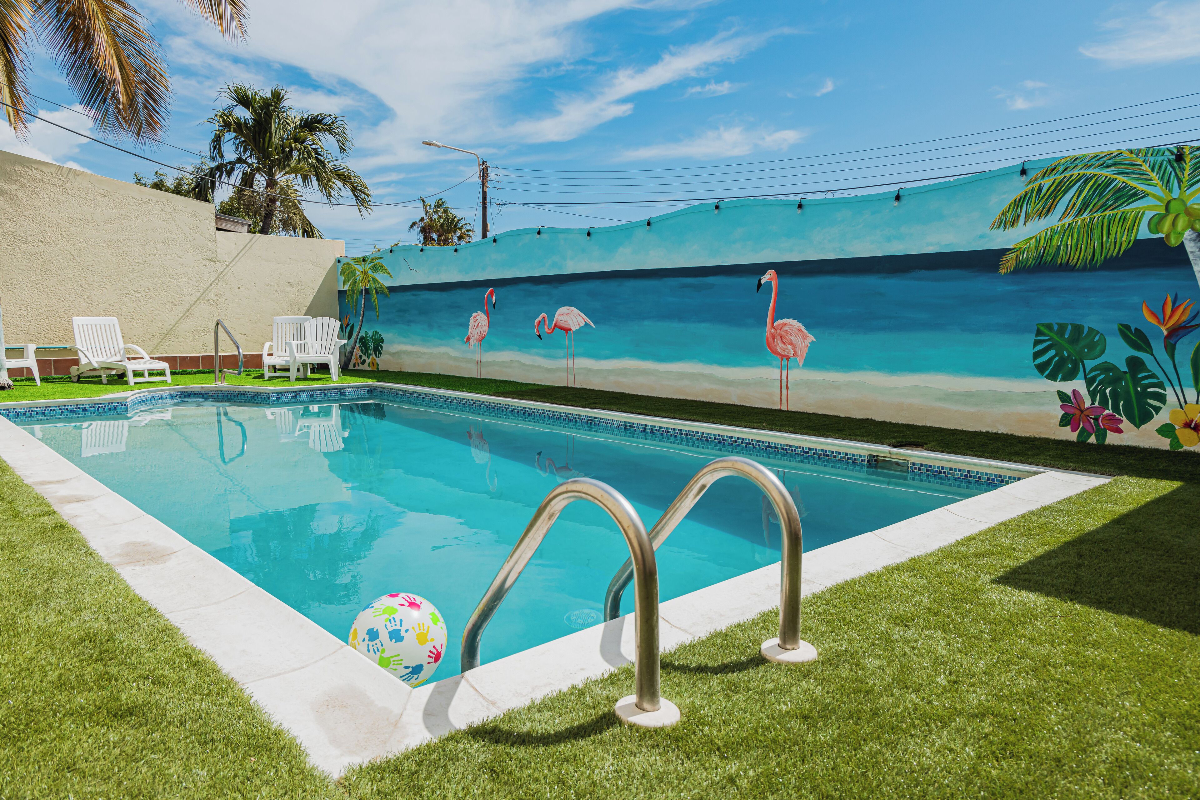 A swimming pool with a mural of flamingos and palm trees, surrounded by white chairs and a grassy area.