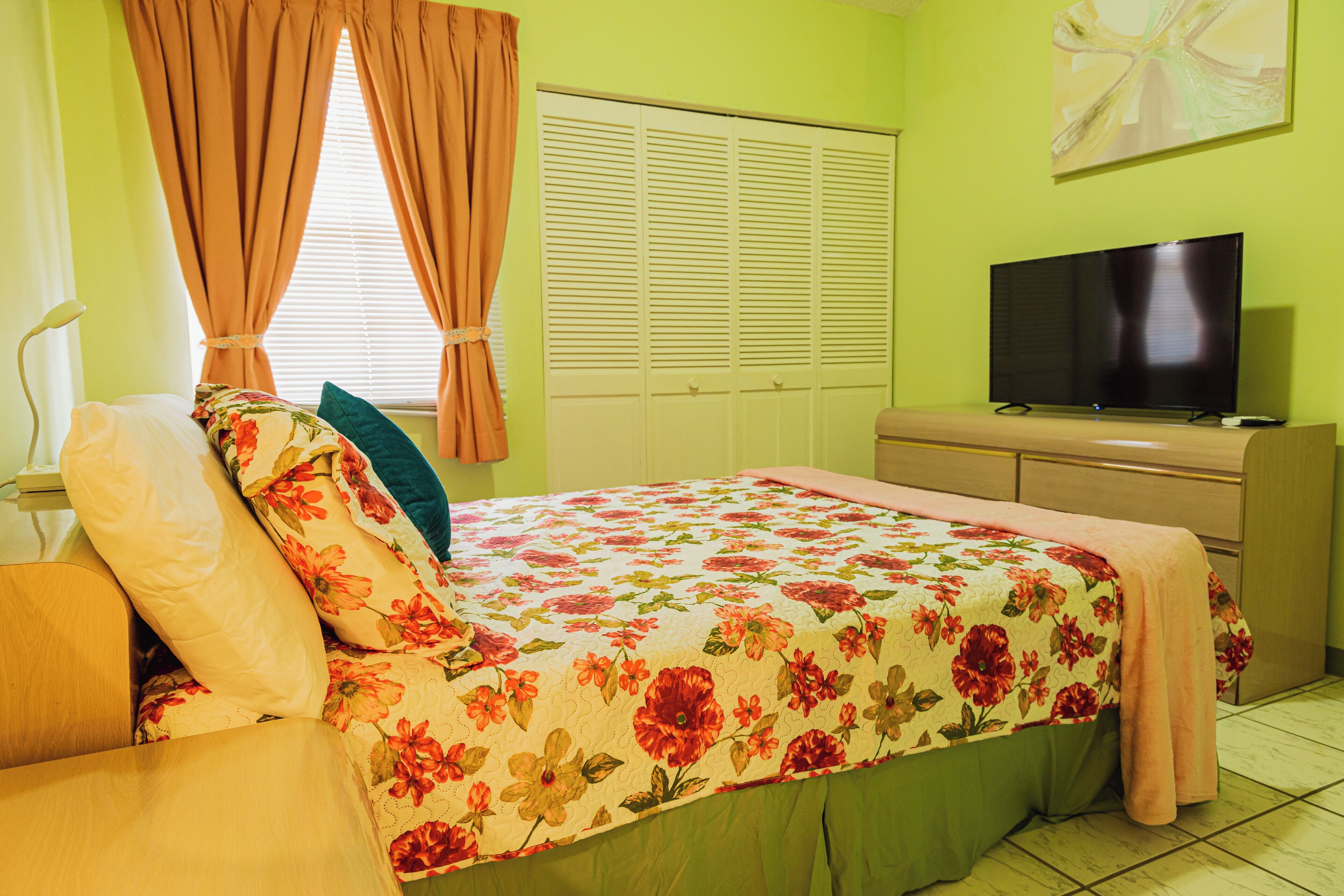 Ein Schlafzimmer mit blumigem Bettüberwurf, einem Fernseher auf einer Kommode und einem Fenster mit Vorhängen.