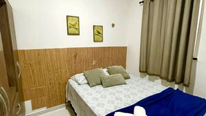 Une chambre d’hôtel avec une tête de lit en bois, un lit recouvert d’une couverture bleue et agrémenté de serviettes blanches, deux tableaux encadrés au mur et une fenêtre avec des rideaux.
