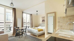 A hotel room with a bed, a desk, a chair, a dining table, a kitchen area with a sink, and a balcony with a view.