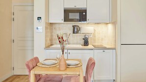 A compact kitchen with white cabinets, a microwave, a kettle, and a table set for two with pink chairs.