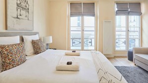 A hotel room with a large bed, two bedside tables, a chair, and a view of the city through large windows.
