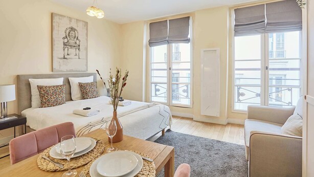 A hotel room with a bed, a dining table set with plates and a glass, a sofa, and large windows with curtains.