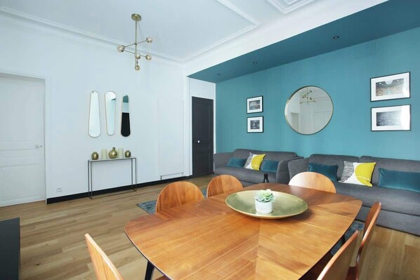 Un salon moderne avec un coin repas, agrémenté d’un mur d’accent bleu, d’un miroir rond et d’un canapé gris avec des coussins.