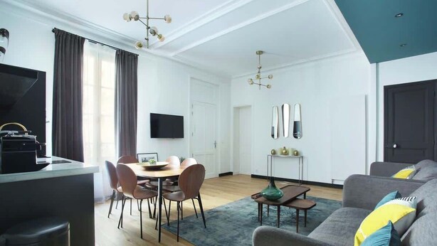A modern living room with a grey sofa, a dining table with chairs, a TV, and a kitchen area with a coffee machine.