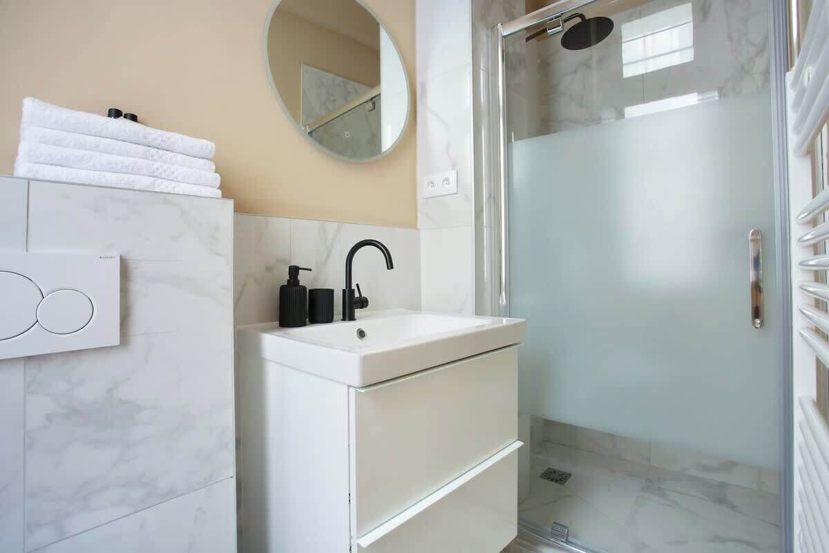 Un bany modern amb un lavabo blanc, un mirall rodó i una dutxa envidriada.