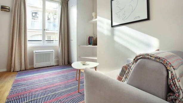 A modern room with a large window, a radiator, a patterned rug, a white sofa, a small white table, and a framed artwork on the wall. - Sublime and new Studio Le Marais / Beaubourg (Paris)