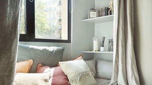 Ein gemütliches Schlafzimmer mit Bett, Kissen, einem Regal mit Büchern und Dekoration sowie einem Fenster mit Blick auf Gebäude.