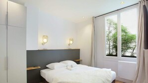 A modern bedroom with a large bed, built-in wardrobe, bedside lamps, and a view of trees through the window. - Elegant Apartment at the Palais Royal Opera (Paris)