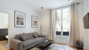 A modern living room with a grey sofa, a black coffee table, and a large window with curtains. - Elegant Apartment at the Palais Royal Opera (Paris)