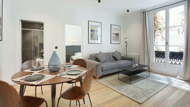 A modern living room with a dining area, a grey sofa, and a coffee table. - Elegant Apartment at the Palais Royal Opera (Paris)