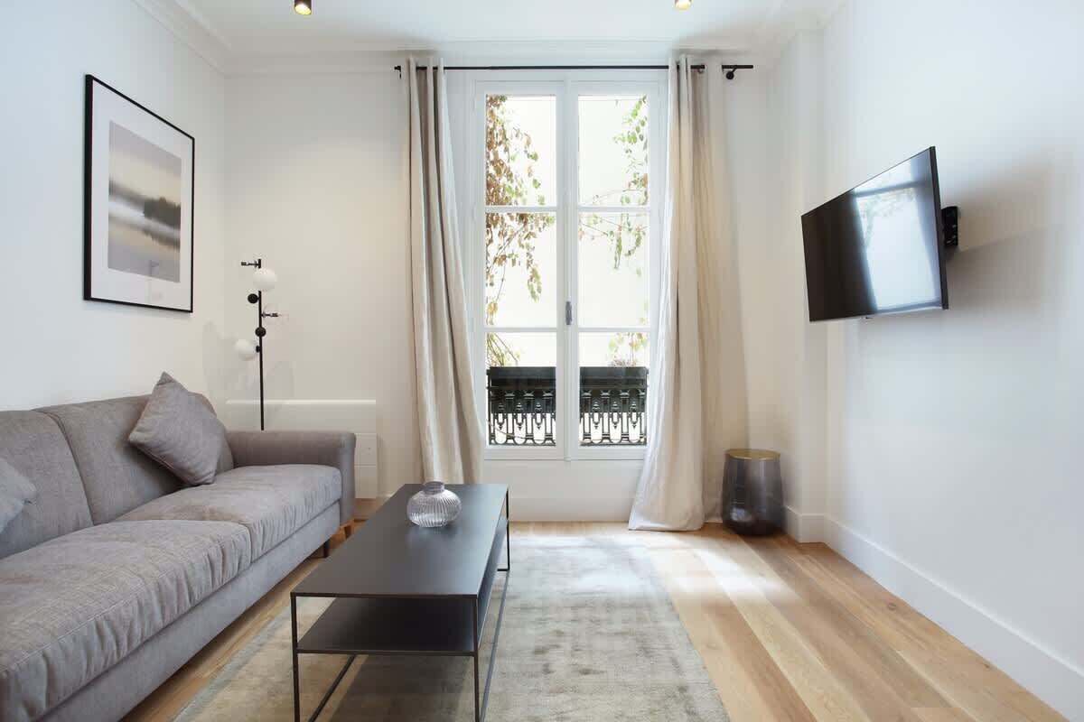 Ein modernes Wohnzimmer mit grauem Sofa, einem schwarzen Couchtisch, einem gerahmten Bild an der Wand, einem Fenster mit Vorhängen und einem an der Wand befestigten Fernseher.