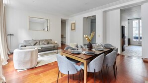 Un comedor moderno con una mesa de madera dispuesta para seis personas, rodeada de sillas tapizadas en gris. En el fondo, un sofá y un sillón blanco pequeño, con un espejo grande en la pared.
