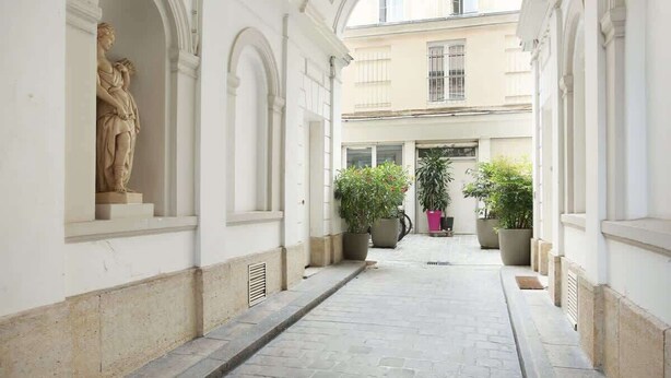 Un estrecho pasillo empedrado de piedra con paredes blancas y arcos, que conduce a un edificio con ventanas y macetas.