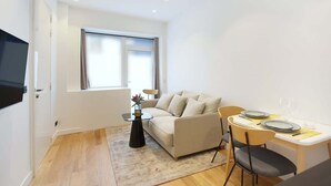 Un espacio habitable compacto con un sofá, una mesa de comedor para dos personas, una mesita auxiliar y un televisor montado en la pared.