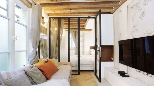 Un dormitorio moderno con techo de madera, una cama con muchas almohadas, un televisor en la pared y un ventanal corredero que da a otra habitación.