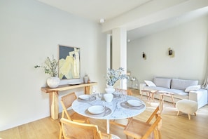 A modern living room with a round dining table set for four, surrounded by wooden chairs. A sofa and a coffee table are visible in the background.
