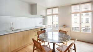 A modern kitchen with a round dining table and wooden chairs, large windows with blinds, and a view of buildings outside. - Sublime Apartment AC Paris  Center (Paris)
