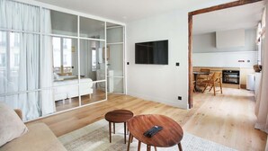 A modern living room with a sofa, two wooden side tables, a TV, and a glass partition leading to a bedroom. - Sublime Apartment AC Paris  Center (Paris)