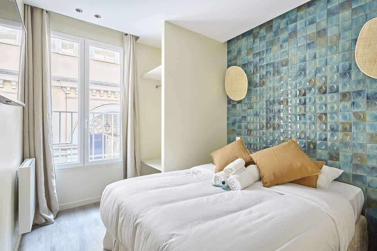 Une chambre d’hôtel avec un grand lit, une fenêtre avec des rideaux, un mur orné d’une mosaïque et un luminaire rond.