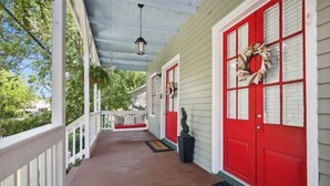 Un porche abrité avec une porte rouge et une guirlande, une balustrade blanche et un banc.
