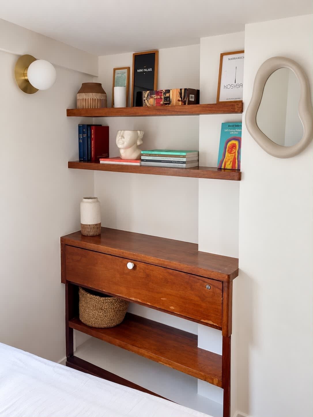 Ein Schlafzimmer mit einem Bett, einem Nachttisch aus Holz mit einer Schublade sowie zwei oberhalb angebrachten Regalen mit Büchern, einer Vase und Dekoration.