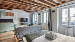 Una habitación de hotel moderna con vigas de madera a la vista, una cama grande con un sofá gris, una kitchenette pequeña y vistas a la ciudad.