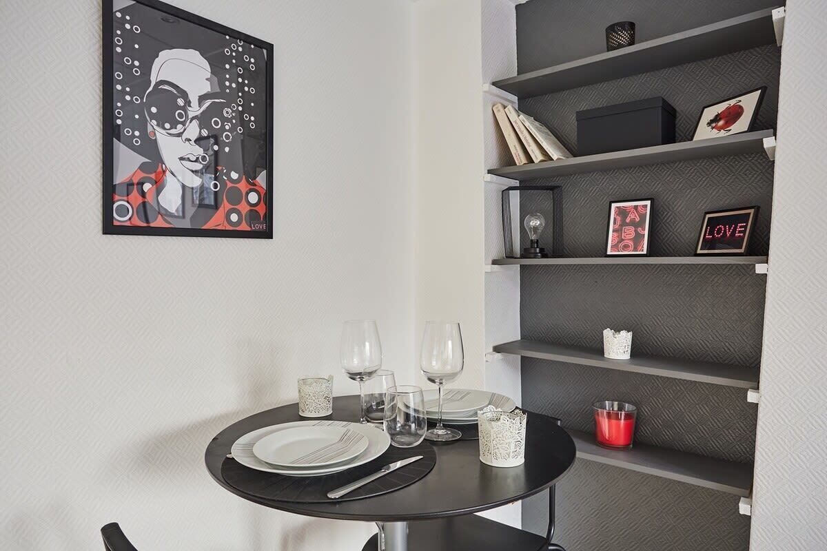 Un área de comedor con una mesa redonda preparada para cuatro personas, una estantería con diversos objetos y una obra de arte enmarcada en la pared.
