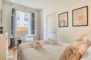 A hotel room with a bed, a chair, a desk, a window with curtains, and artwork on the wall. - Eiffel Tower Terrace Apartment (Paris)